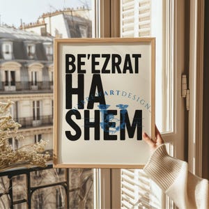 Può includere: Una stampa incorniciata con le parole "BE'EZRAT HA SHEM" in nero, con un disegno floreale blu. La stampa è tenuta davanti a una finestra con vista su un edificio. La cornice è di colore marrone chiaro.