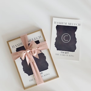Può includere: Una stampa incorniciata con un design in bianco e nero di un pezzo di carta strappato con il testo "Hashem Melech" e "The Lord is King" in ebraico e inglese. La stampa è avvolta in una scatola regalo con un nastro rosa.