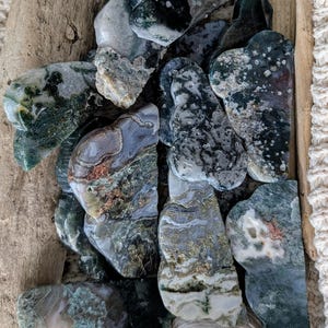 May include: A collection of polished moss agate stones in shades of green, white, and gray. The stones have unique patterns and shapes, with some resembling organic forms. They are arranged on a wooden surface.