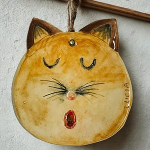 Può includere: Una decorazione da parete rotonda in ceramica smaltata a forma di muso di gatto con finitura beige e marrone. Il gatto ha gli occhi chiusi, baffi dipinti, un piccolo naso rosa e la bocca aperta. Il nome LUCIA è inciso sul lato destro dell'oggetto.