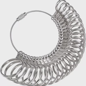 May include: A set of silver-colored ring sizers, connected by a metal cable. Each ring is labeled with a size number, ranging from 0 to 13. The sizers are arranged in a semi-circular shape, ideal for measuring ring sizes.