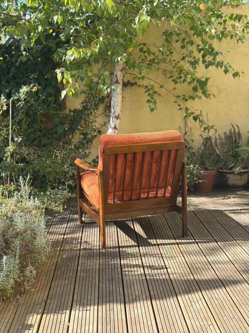 Pu&ograve; includere: Una poltrona in velluto a coste arancione con struttura in legno. La sedia &egrave; posizionata su un terrazzo in legno, con un albero e vegetazione sullo sfondo. Le dimensioni della sedia sono di circa 76 cm di larghezza, 81 cm di profondit&agrave; e 86 cm di altezza.