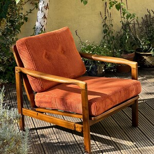 Pu&ograve; includere: Poltrona vintage con cuscino e schienale in velluto a coste arancione. La struttura in legno &egrave; di colore marrone caldo. La sedia &egrave; all'aperto, sullo sfondo di piante e un albero. Il design della sedia ricorda i mobili della met&agrave; del secolo.