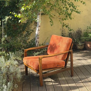 Pu&ograve; includere: Una poltrona in velluto a coste arancione con struttura in legno. La sedia &egrave; su un terrazzo in legno. La sedia ha schienale alto e braccioli. La sedia &egrave; in un giardino con un albero e piante sullo sfondo.