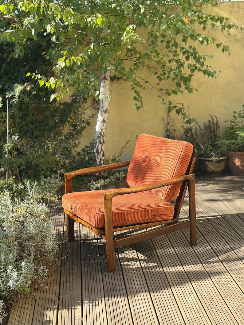 Pu&ograve; includere: Una poltrona in velluto a coste arancione con struttura in legno. La sedia &egrave; su un terrazzo in legno. La sedia ha schienale alto e braccioli. La sedia &egrave; in un giardino con un albero e piante sullo sfondo.
