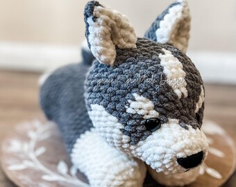 May include: A crocheted husky puppy toy with gray and white fur, lying on a wooden plate with a floral design.