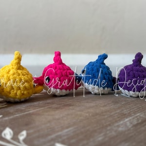 May include: A collection of six crocheted shark toys in various colors: orange, yellow, pink, blue, purple, and green. Each shark has a white belly and black eyes. They are arranged on a wooden decorative tray.