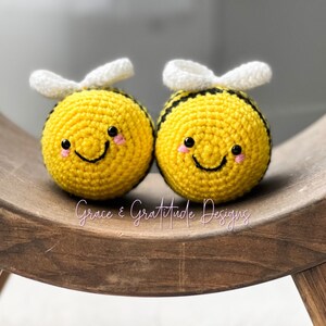 May include: Two crocheted bumblebees with black and white striped bodies, yellow heads, and white wings. The bumblebees have black eyes and pink cheeks and are smiling. The bumblebees are sitting on a wooden surface.