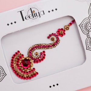 May include: A decorative bindi sticker featuring a swirling design of pink and gold rhinestones with a large central pink gem. The adhesive accessory is displayed inside a white rectangular cardboard package labeled with the brand name Teeliya.