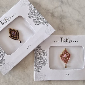 May include: Two bindi sets in white rectangular packaging with mandala-style corner prints and the text Teeliya. One bindi is diamond-shaped with dark brown stones and a central purple gem, while the other features pink stones and a central pink gem.