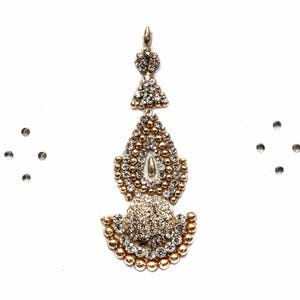May include: A gold-toned maang tikka headpiece featuring a central teardrop-shaped pendant adorned with clear crystals and small gold beads. The piece includes a top floral accent and a semi-circular base, accompanied by two separate four-stone clusters.