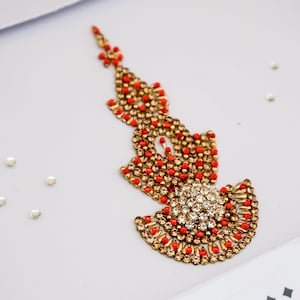 May include: A decorative forehead ornament featuring a vertical, pointed design adorned with small, metallic gold-toned beads, vibrant orange accents, and a central circular cluster of clear, sparkling rhinestones, accompanied by four loose rhinestones.