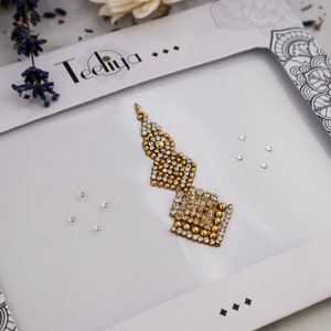 May include: A gold-toned bindi with a geometric, multi-tiered design featuring clear crystals and small gold beads. The bindi is displayed on a white card labeled Teeliya, accompanied by several loose, individual clear crystal accents.