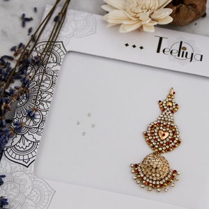 May include: A gold-toned decorative bindi or forehead ornament featuring a central heart shape surrounded by small clear and red rhinestones. The piece is attached to a white display card labeled Teeliya with black mandala-style line art.
