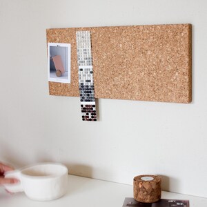 May include: A corkboard with a light brown finish, mounted on a white wall. A small photo and a sample tile sheet are pinned to the corkboard.
