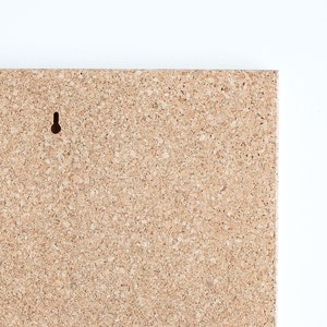May include: A close-up of a corkboard with a small hole in the top left corner. The corkboard is a light brown color and has a rough texture.