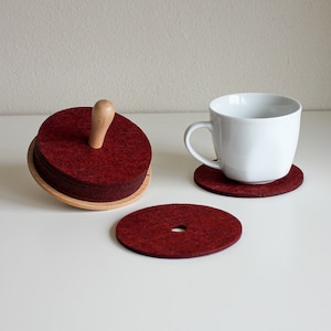 Può includere: Un set di sottobicchieri rotondi in feltro bordeaux, con supporto e manico in legno. Una tazza bianca poggia su un sottobicchiere abbinato. Un sottobicchiere è forato.