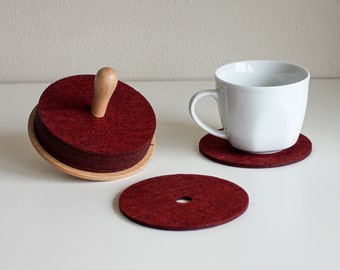 May include: A set of burgundy felt coasters with a wooden holder and handle. A white mug sits on a matching coaster. The coasters are round and have a hole in the center of one.