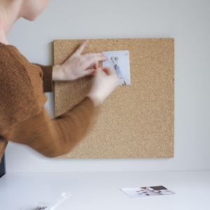 May include: A person pinning a photo to a corkboard. The photo shows a person's legs and feet in the air. The corkboard is brown and has a rough texture.