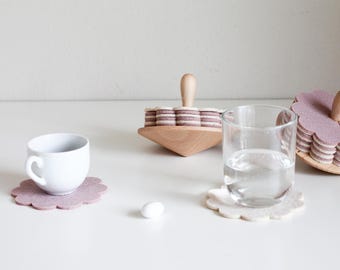 May include: A white teacup and a glass of water sit on a white surface, next to a light purple felt coaster. A wooden coaster holder with stacked felt coasters and a wooden handle is also present. The coasters are flower-shaped.