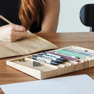 May include: A wooden organizer with multiple slots for holding pens, pencils, and markers. The organizer is made of light wood and has a natural finish. The organizer is sitting on a table with a white piece of paper in front of it.