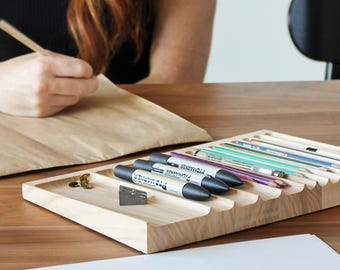 Puede incluir: Un organizador de madera con múltiples ranuras para guardar bolígrafos, lápices y rotuladores. El organizador está hecho de madera clara y tiene un acabado natural. El organizador está sentado en una mesa con una hoja de papel blanca delante.