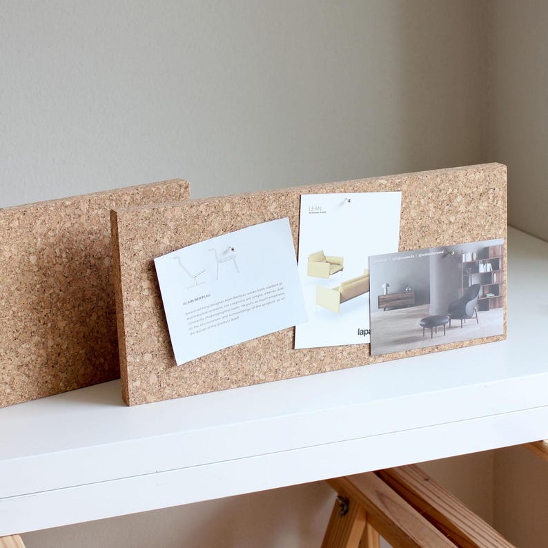May include: Two cork boards with a light brown color, standing upright on a white surface. The boards are covered with cork and have several pieces of paper pinned to them. The papers contain images and text about furniture design.