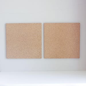 May include: Two cork boards, each with a light brown color, are mounted on a white wall. The boards are square shaped and have a textured surface.