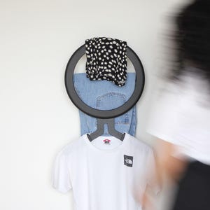 May include: A circular black wall-mounted clothing rack holding a pair of blue denim jeans, a black and white floral patterned garment, and a white short-sleeved t-shirt with a small black square logo on the chest area of the fabric.