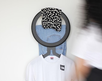 May include: A circular black wall-mounted clothing rack holding a pair of blue denim jeans, a black and white floral patterned garment, and a white short-sleeved t-shirt with a small black square logo on the chest area of the fabric.