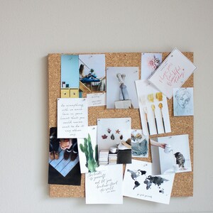 May include: A corkboard with various inspirational quotes, photos, and artwork. The corkboard is covered in a brown cork material and has a variety of push pins holding the items in place. The items include a quote about love, a photo of a woman in jeans, a photo of a forest, and a photo of a world map.
