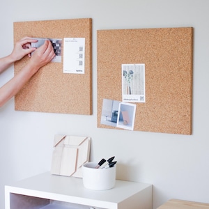 May include: Two cork boards with different sizes and shapes, both with printed papers and photos pinned to them. The boards are mounted on a white wall. A white shelf with a wooden object and a white cup with black markers is below the boards.