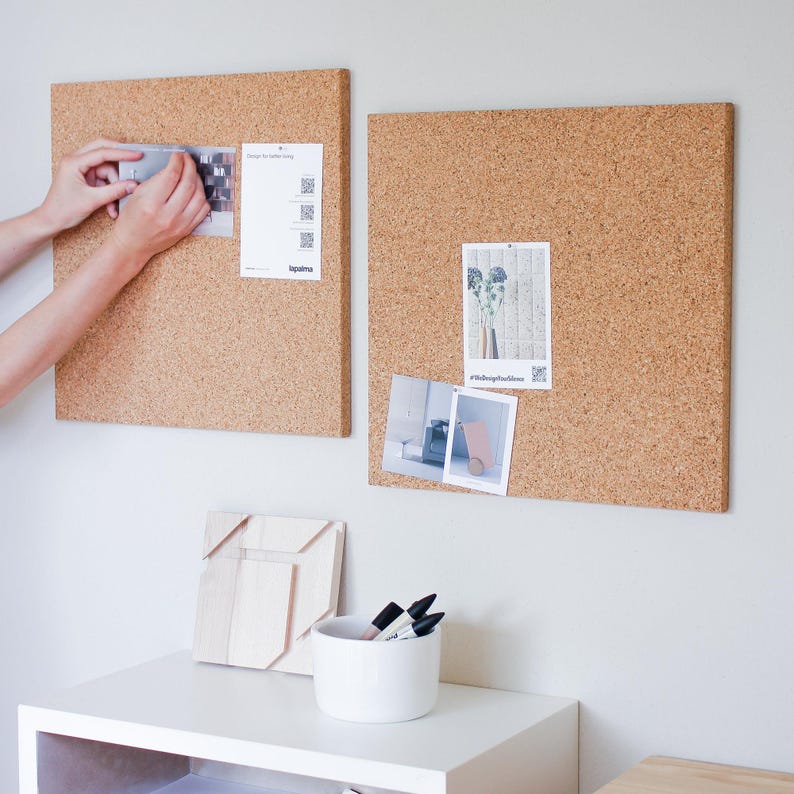 May include: Two cork boards with different sizes and shapes, both with printed papers and photos pinned to them. The boards are mounted on a white wall. A white shelf with a wooden object and a white cup with black markers is below the boards.