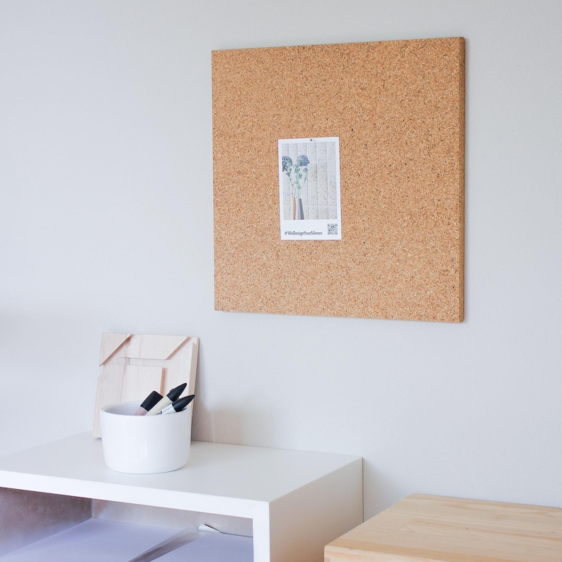 May include: A corkboard with a photo of a plant pinned to it. The corkboard is mounted on a white wall. There is a white shelf with a white bowl and black markers on it.