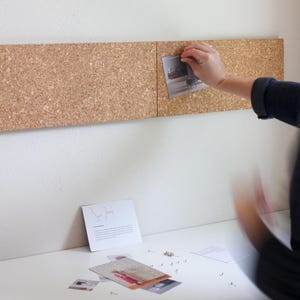 May include: A cork board with a light brown color, mounted on a white wall. A person is pinning a piece of paper with a design on it to the board. There are also some small pieces of paper and push pins on a white table below the board.