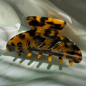 May include: A brown and black tortoiseshell hair claw with the name "Natalie" engraved in gold lettering.