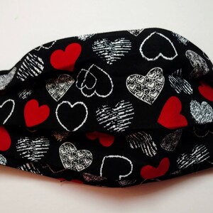 May include: A black face mask with a pattern of red, white, and black hearts. The mask has white elastic ear loops. The hearts are in various styles, including solid red, white outlines, and textured designs. The mask is pleated for a comfortable fit.