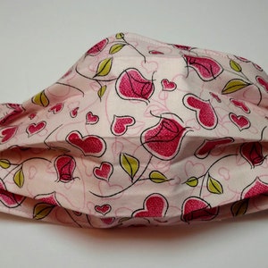 May include: A pink face mask with a white background and a pattern of pink hearts and green leaves.