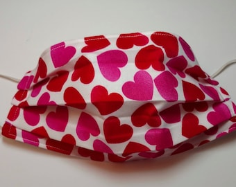 May include: A white face mask with a pink and red heart pattern. The mask has elastic ear loops.