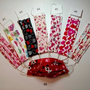 May include: A collection of eight face masks with various patterns. Designs include hearts, floral, and a dog with the text "I woof you". Each mask has white elastic ear loops. The masks are numbered from 1 to 8.