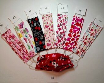 May include: A collection of eight face masks with various patterns. Designs include hearts, floral, and a dog with the text "I woof you". Each mask has white elastic ear loops. The masks are numbered from 1 to 8.