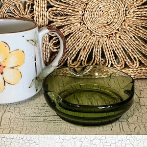 May include: A dark green glass ashtray with a rounded, slightly heart-shaped design. The ashtray has a smooth, glossy finish.