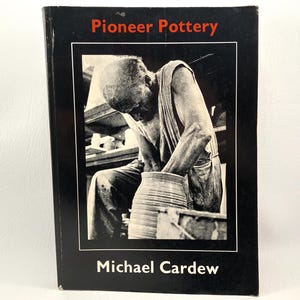 May include: A book titled "Pioneer Pottery" with a black cover and a black and white image of a potter working on a pot. The author's name, Michael Cardew, is printed at the bottom of the book.