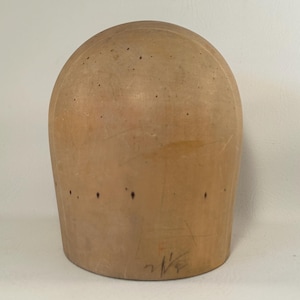 May include: A vintage hat block made of light brown wood. The hat block has a rounded top and a cylindrical base. It has several small holes and markings on the surface. The hat block is used for shaping hats.