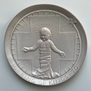 May include: A round, light gray ceramic plate with a raised relief design. The design features a baby with a halo, a cross, and a star. The words "19-KING OF KINGS-70" are inscribed around the edge of the plate. The rim of the plate is a darker shade of gray.