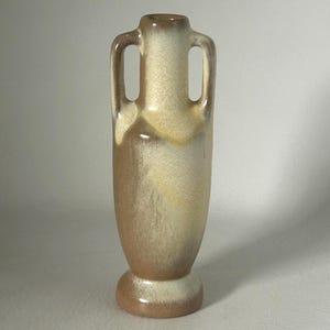 May include: A tall ceramic vase with two integrated handles, featuring a mottled glaze in shades of cream, beige, and brown. The vessel has a narrow neck, a rounded body, and a circular base, with a smooth, blended color transition throughout.