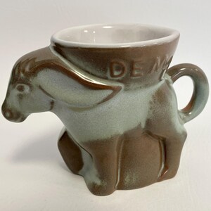 May include: A ceramic donkey-shaped mug with a handle. The mug is primarily brown with light blue and white accents. The word "DEM" is visible on the side of the mug.