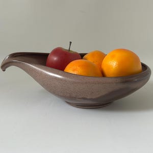 May include: A brown ceramic fruit bowl with a unique, curved design. The bowl contains a red apple and several oranges. The bowl has a speckled texture and a smooth, glossy finish. The fruit is fresh and vibrant.