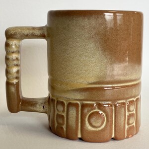 May include: A brown ceramic mug with a textured handle and a decorative base. The mug has a cylindrical shape with a gradient of colors, transitioning from light beige to a darker brown. The handle has a ridged design.