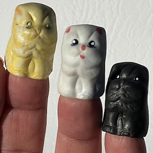 May include: Three finger puppets shaped like cats. The first cat is yellow, the second is white with pink accents, and the third is black.